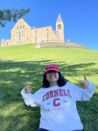 Maria Delgado stands at the bottom of Libe Slope. She's a young woman, pointing at the Cornell hoodie she's wearing while the clocktower sits on the hill behind her. 