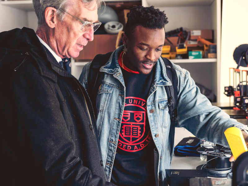 A Cornell student wearing a sweatshirt with the Cornell seal gestures and shows Cornell alumnus Bill Nye something off image.