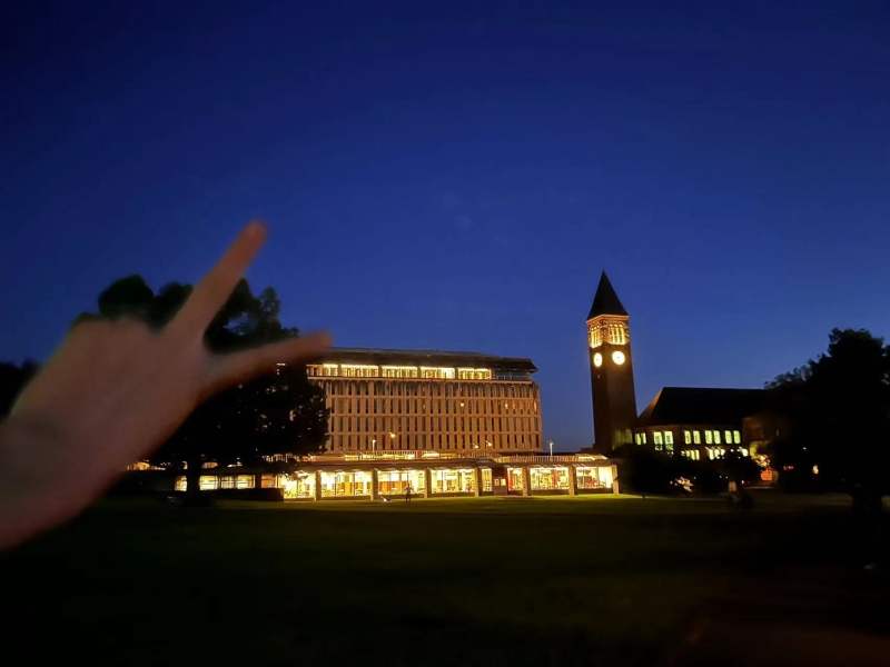 The photo taker takes a picture of Olin Library lit up at night with their hand in the picture making a peace sign.