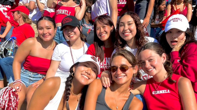 Eight Cornell students gather in Cornell red in the bleachers at the Homecoming football game smiling widely.